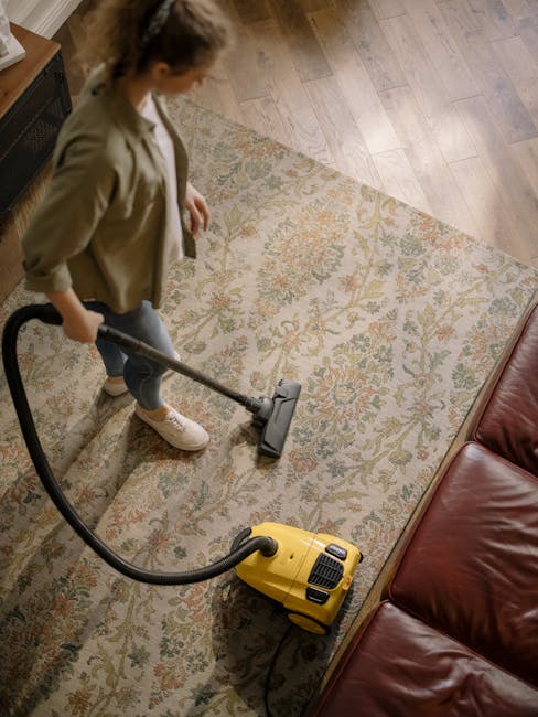 A professional cleaning specialist dressed in white protective coveralls, gloves, and a face mask is conducting deep carpet cleaning in a bright, modern living room. The individual is using a speciali