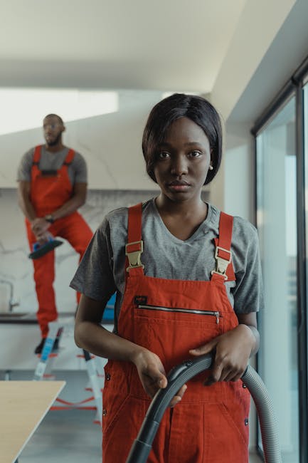 A professional cleaner, a young woman with dark hair, is holding a vacuum cleaner hose in a modern residential interior, focusing on surface cleaning. She is wearing a grey t-shirt and bright orange work overalls, indicating a cleaning service. In the background, another worker in similar attire stands slightly blurred, with a concrete wall and a large window providing natural lighting to the bright, clean space. The room appears to be a living area or open plan space with minimal furnishings, showcasing surfaces like wooden tables and tiled floors that are visibly clean and well-maintained. The emphasis is on thorough domestic cleaning and surface sanitisation, consistent with the services discussed on BarnesCleaner.co.uk for end of tenancy cleaning rates in SW13. The professional cleaning team demonstrates attention to detail and effective hygiene practices within a contemporary home environment.