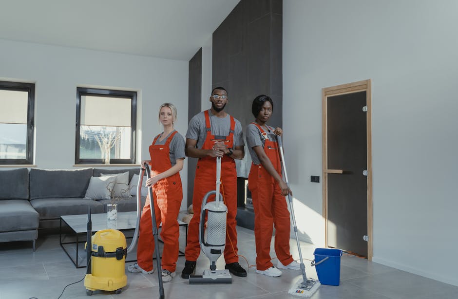 Three professional cleaners wearing grey shirts and red overalls stand in a modern living room, equipped with cleaning tools including a vacuum cleaner, mop, and bucket. They are positioned near a dark-colored door, with a light grey sofa and large windows in the background allowing natural light to illuminate the space. The room features a tiled floor, white walls, and minimal decor, illustrating a clean and well-maintained environment. The image emphasizes surface cleaning and routine domestic cleaning services provided by Barnes Cleaner, highlighting their expertise in deep cleaning and sanitisation in residential spaces.