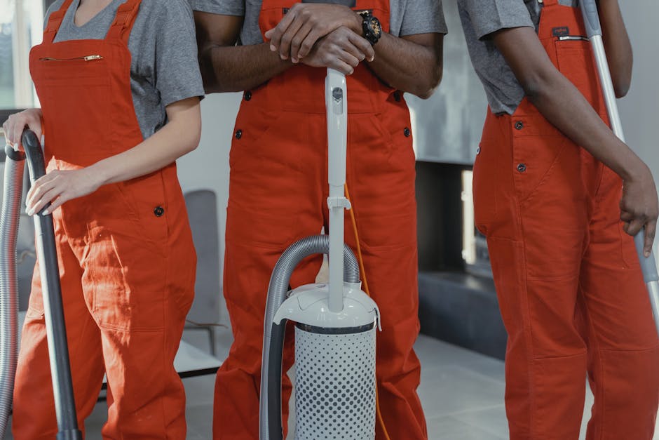 A professional cleaner, a young woman with dark hair, is holding a vacuum cleaner hose in a modern residential interior, focusing on surface cleaning. She is wearing a grey t-shirt and bright orange work overalls, indicating a cleaning service. In the background, another worker in similar attire stands slightly blurred, with a concrete wall and a large window providing natural lighting to the bright, clean space. The room appears to be a living area or open plan space with minimal furnishings, showcasing surfaces like wooden tables and tiled floors that are visibly clean and well-maintained. The emphasis is on thorough domestic cleaning and surface sanitisation, consistent with the services discussed on BarnesCleaner.co.uk for end of tenancy cleaning rates in SW13. The professional cleaning team demonstrates attention to detail and effective hygiene practices within a contemporary home environment.